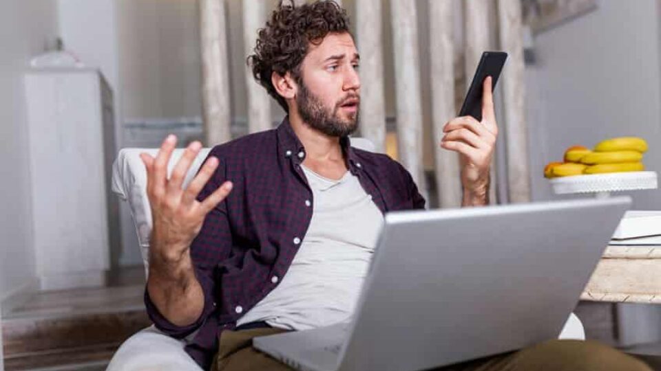 A man despairs in front of his computer and his smartphone, which he holds in his hand.