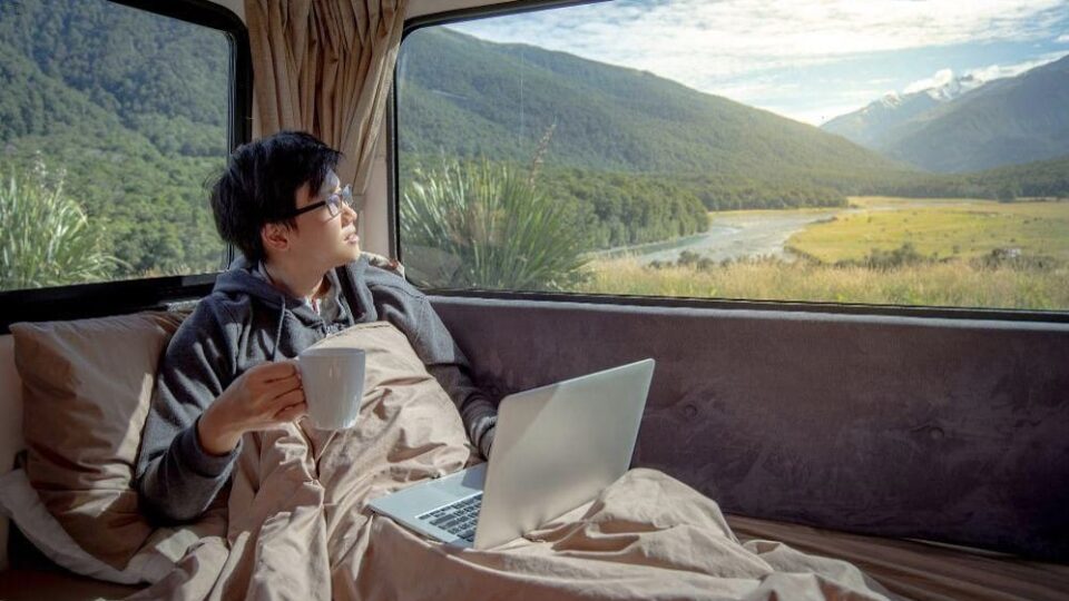 A young man sits in his camper van with a laptop and coffee, looking out at nature.