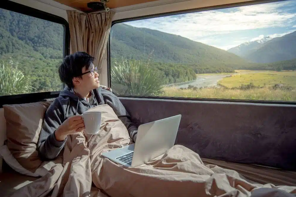 Ein junger Mann sitzt mit Laptop und Kaffee in seinem Camper-Van und schaut in die Natur.