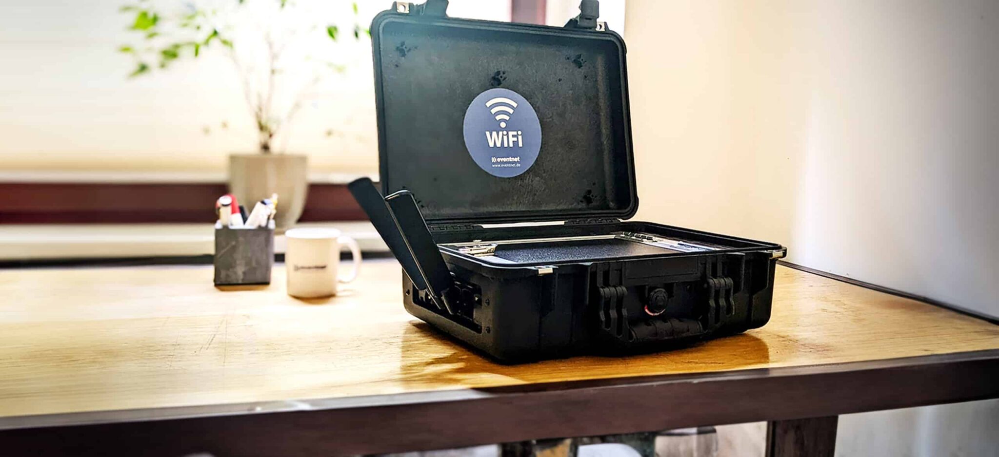 Ein geöffneter, schwarzer Transportkoffer (Hotspot in a Box) mit integriertem WLAN-System steht auf einem Holztisch. Im Kofferdeckel ist ein blaues WiFi-Logo mit der Aufschrift ‚WiFi – Eventnet‘ angebracht. Seitlich ragen zwei Antennen heraus.